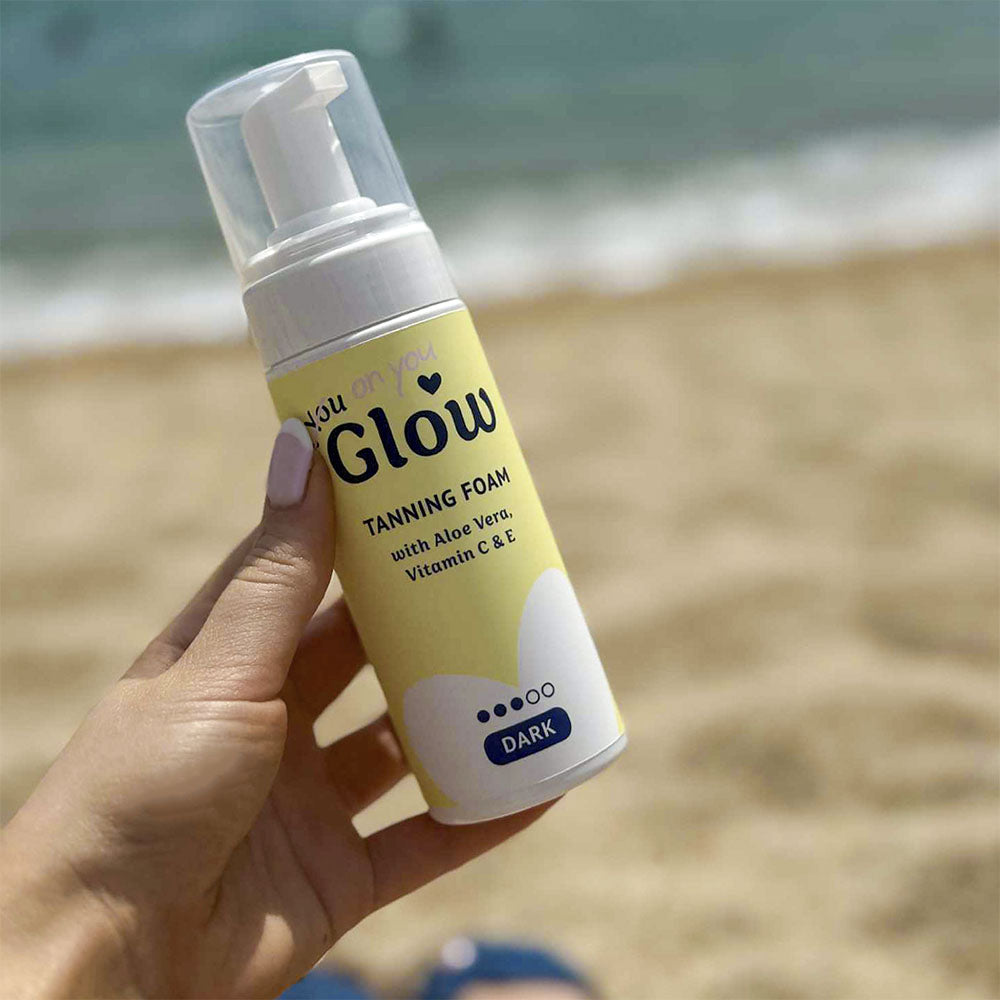 Hand holding a bottle of 'You Glow' tanning foam on a beach.
