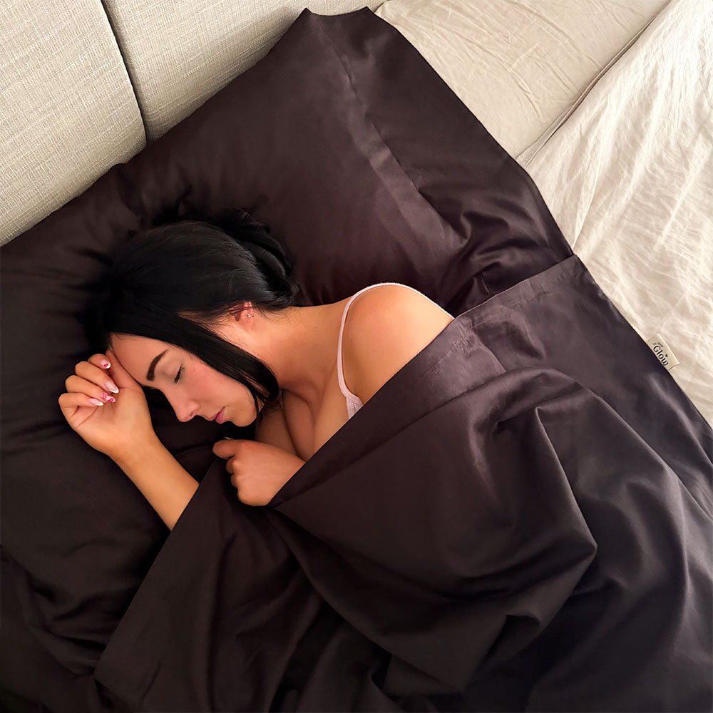 A woman sleeping in a dark brown fake tan sleeping bag, with light beige bedding in the background.