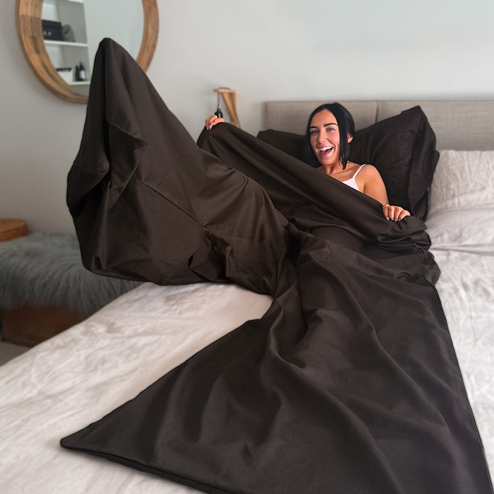 A woman smiling while lying in bed inside a large dark brown self tanning bag, with the fabric spread across the bed in a bedroom setting.